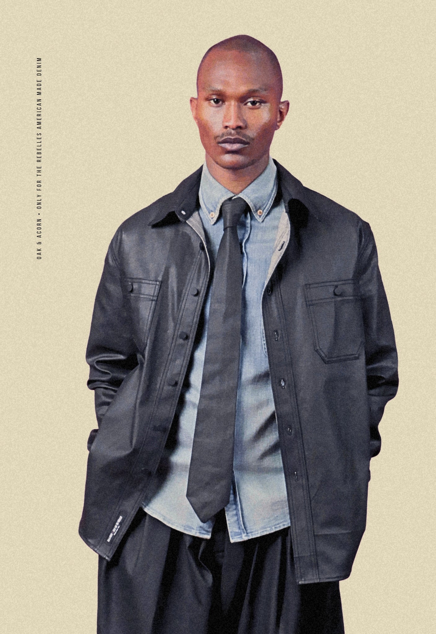 Man wearing a coated black denim chore shacket over a light denim shirt with a coated black denim tie on a beige background