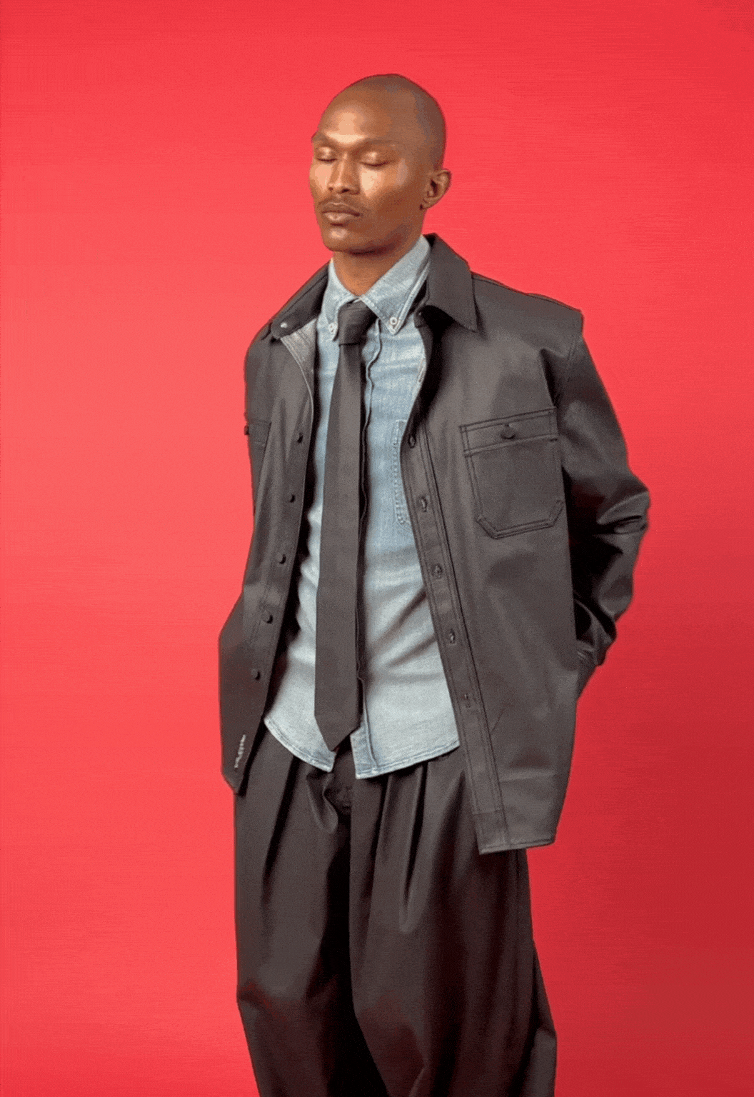 Man wearing a coated black denim chore shacket over a light denim shirt with a coated black denim tie on a red background
