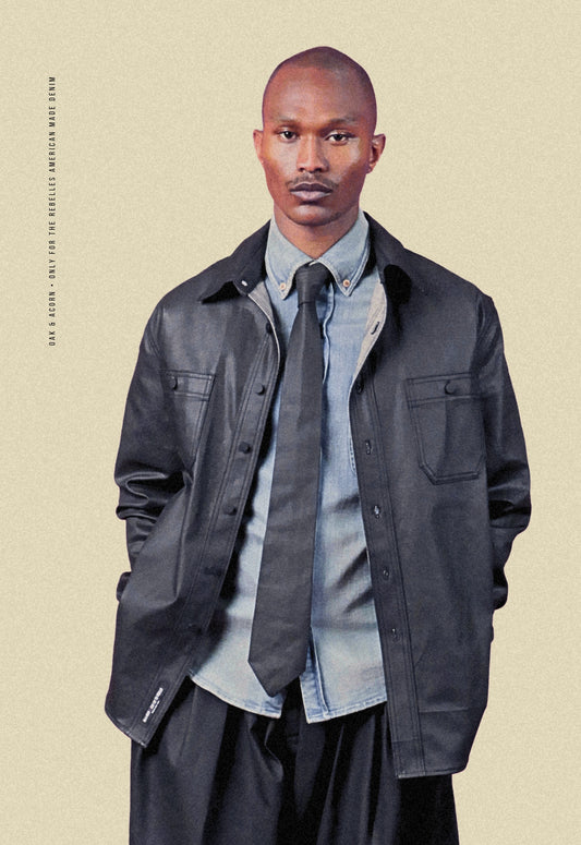 Man wearing a coated black denim chore shacket over a light denim shirt with a coated black denim tie on a beige background