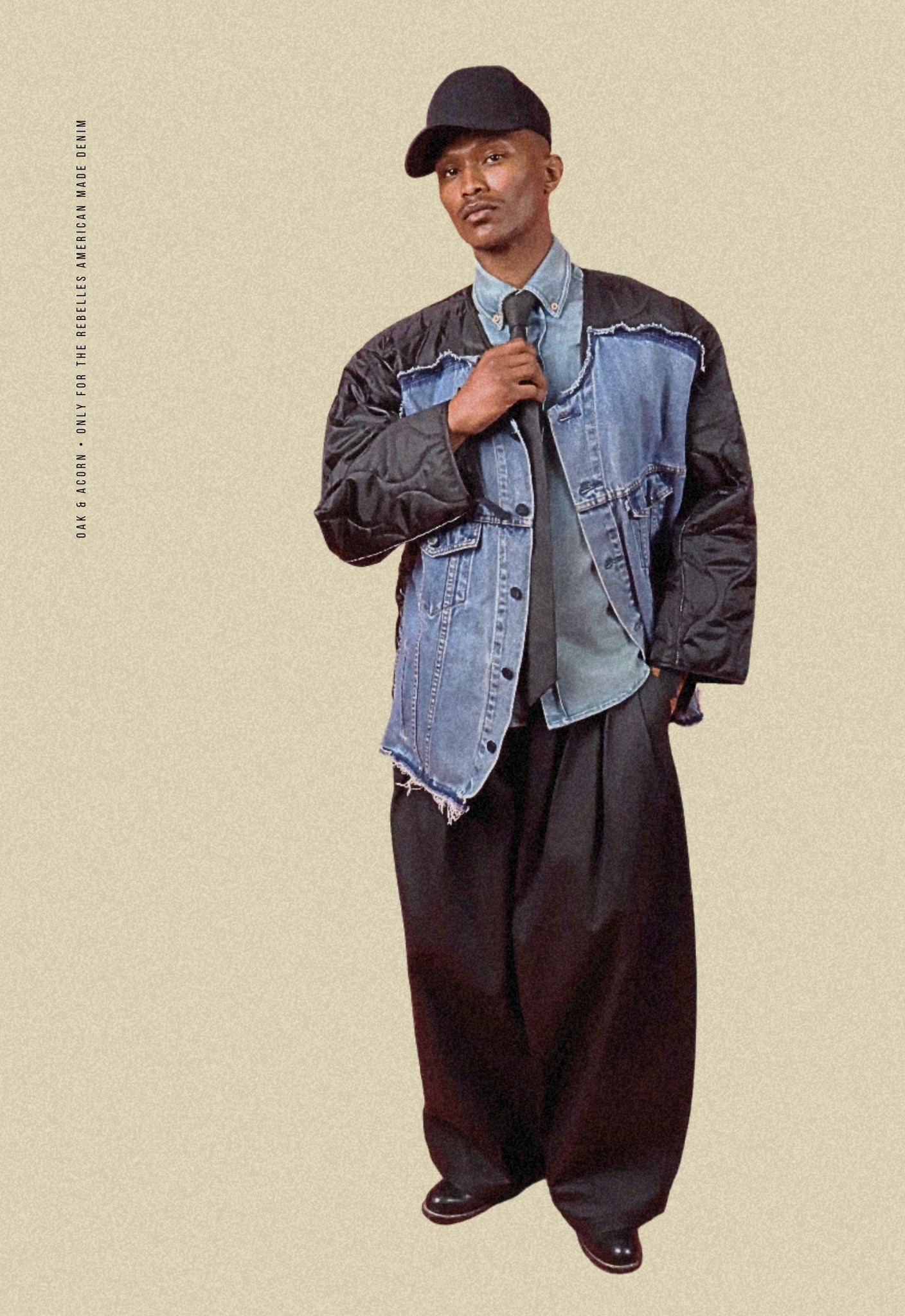 Man wearing coated black denim double waistband wide-leg trousers, repurposed vintage denim bomber, light denim button-down shirt, and coated black denim tie on a beige background