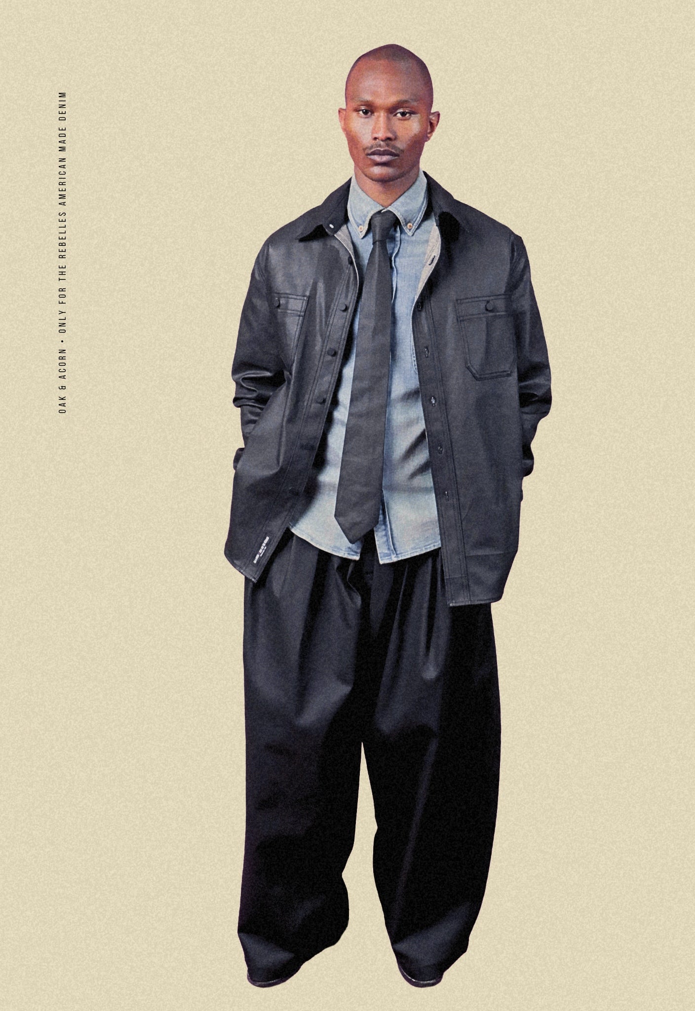 Man wearing coated black denim double waistband wide-leg trousers, coated black denim shacket, light denim button-down shirt, and coated black denim tie on a beige background